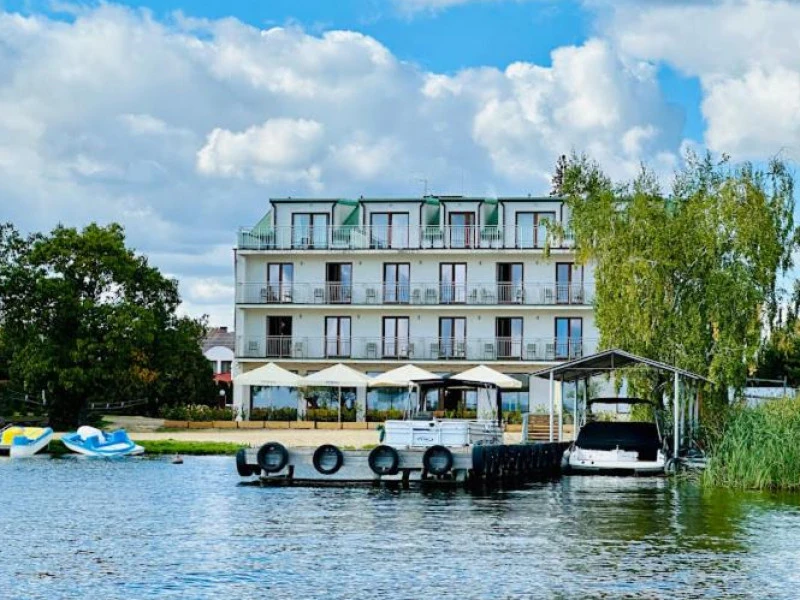 Three-story lakeside hotel with multiple balconies, outdoor seating under white canopies, and a small docked boat at a marina